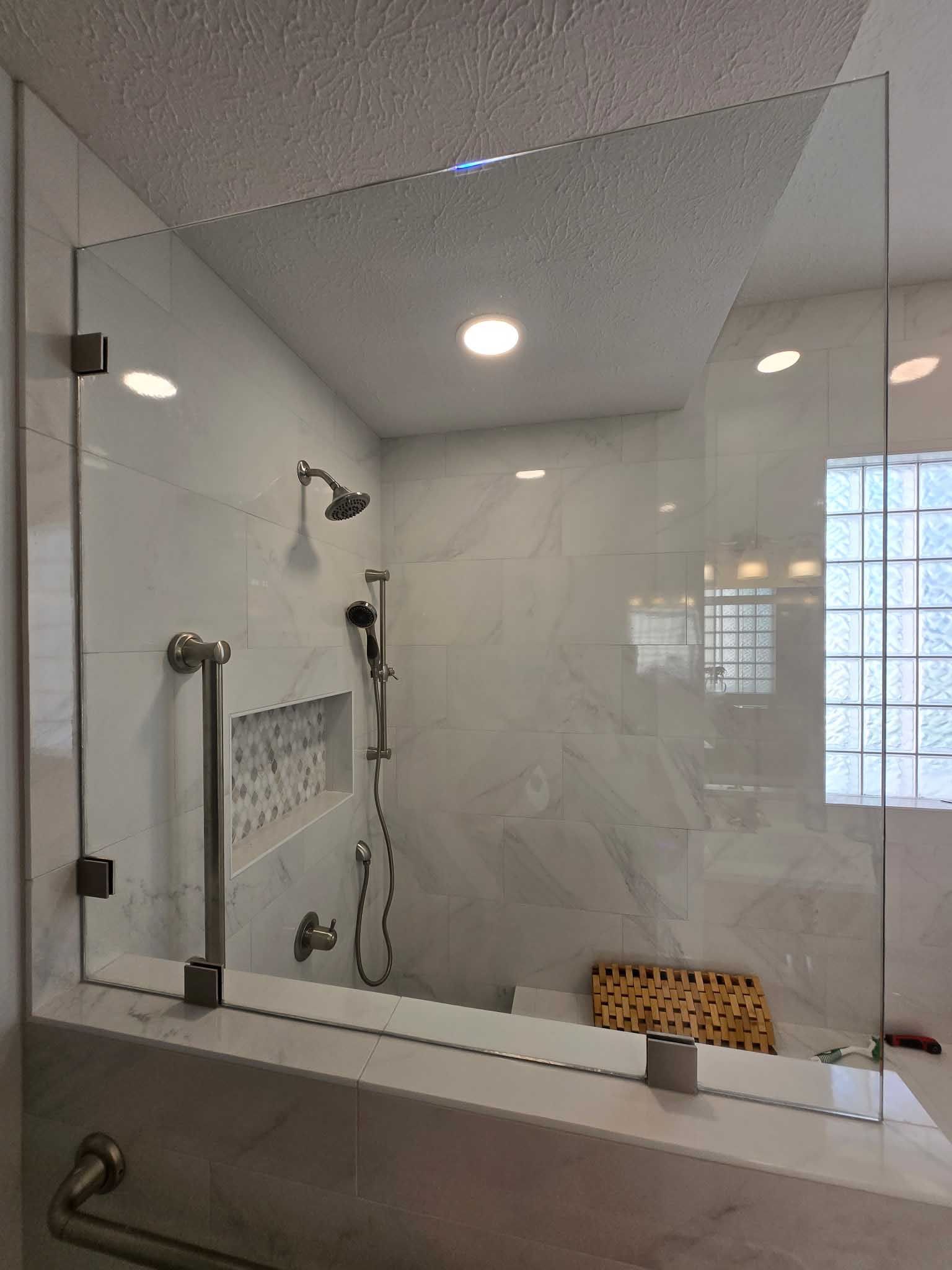 A walk-in shower with white marble-style walls, glass paneling, a handheld showerhead, and a built-in shelf.