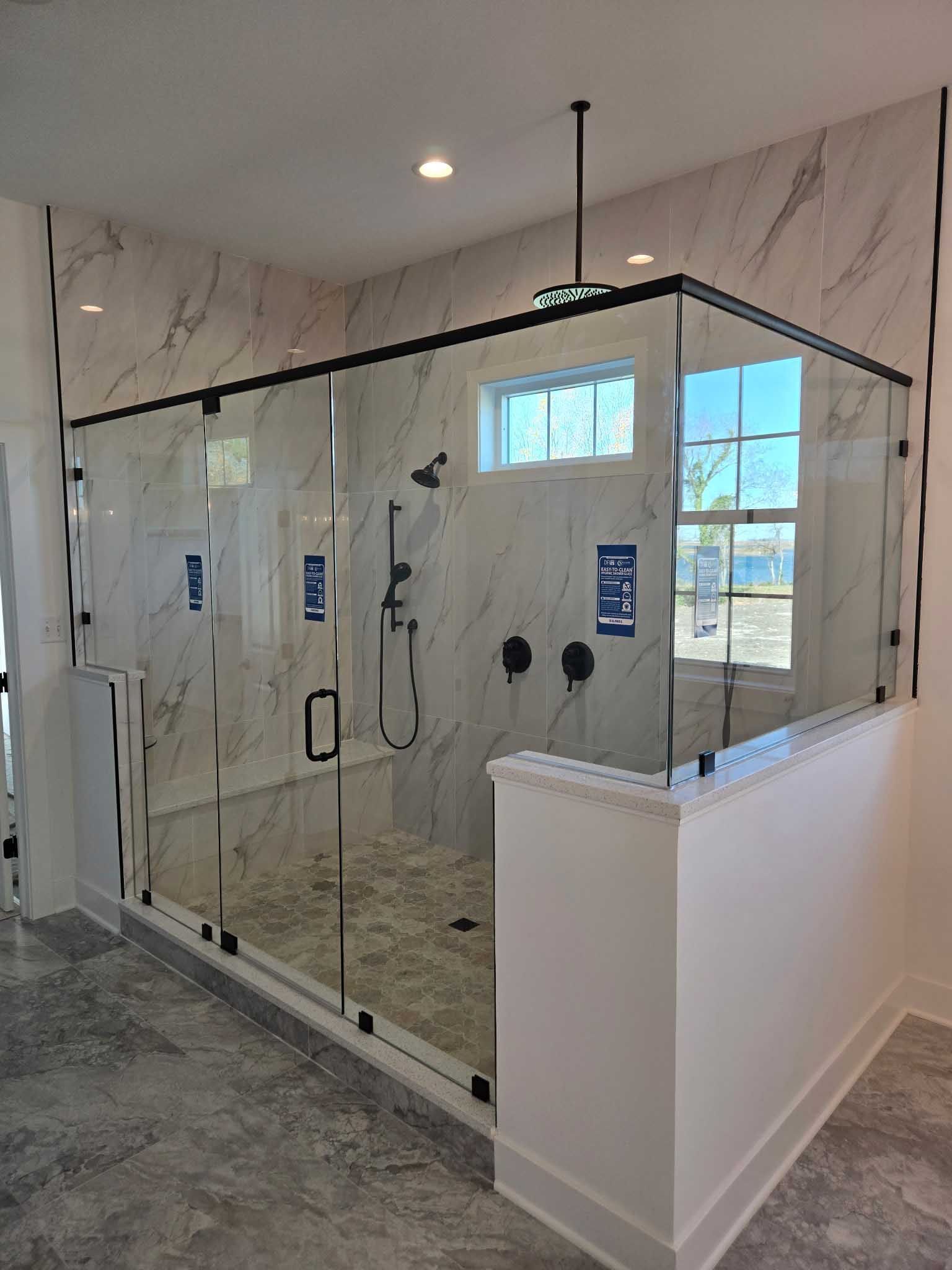 A walk-in shower with glass walls, marble-patterned tile, and a black fixture set in a modern, brightly lit bathroom.