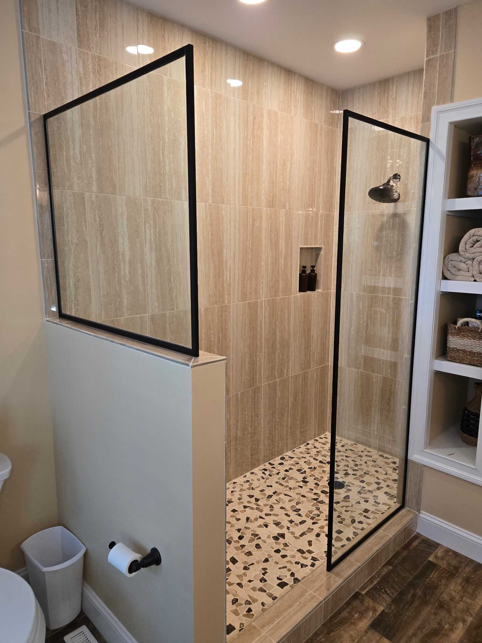 Modern walk-in shower with beige tiled walls, a pebble floor, black-framed glass panels, and an adjacent open shelf unit.