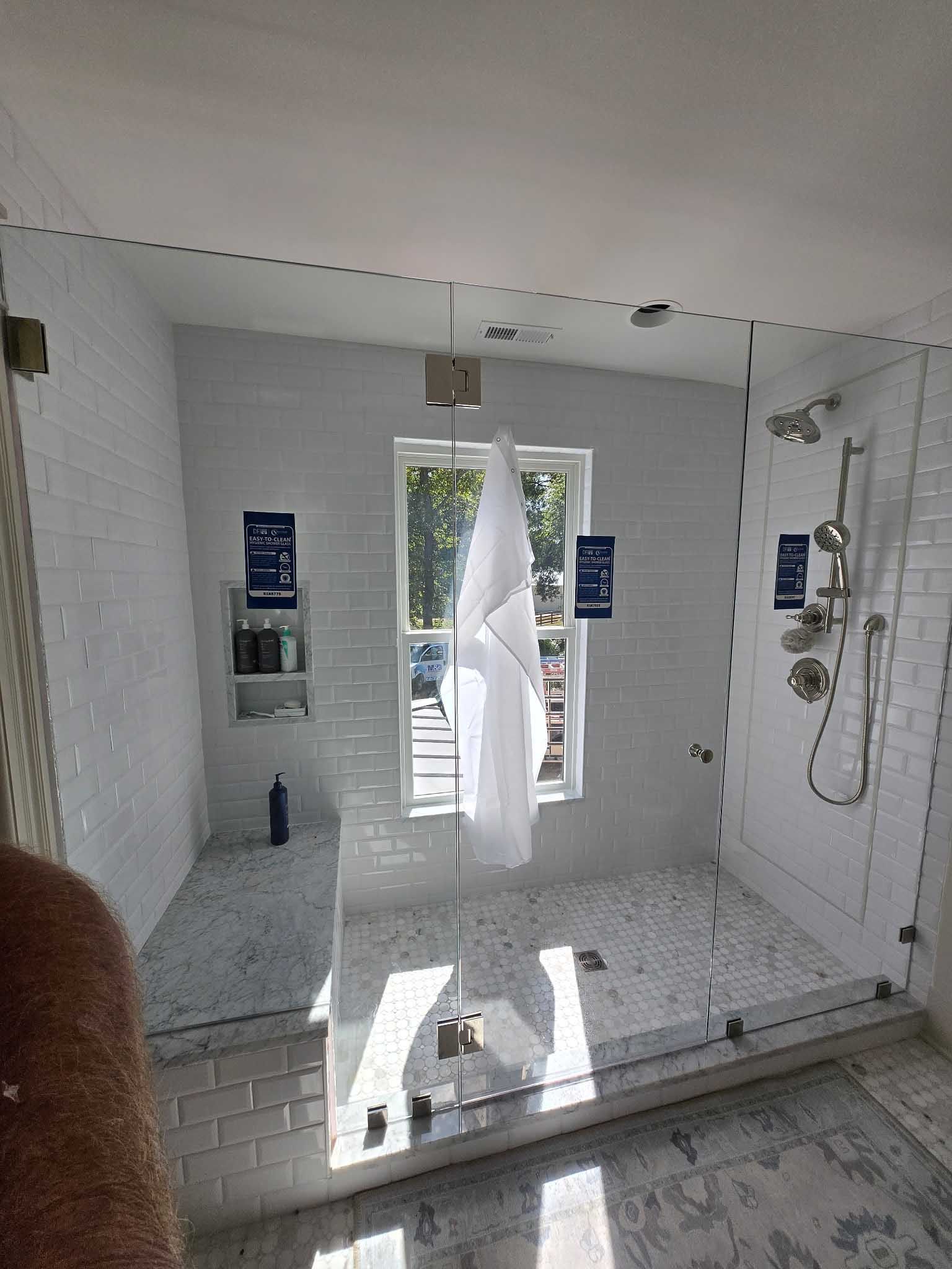 A walk-in shower with white brick walls, marble floor, a built-in bench, and a glass partition in front of a window.
