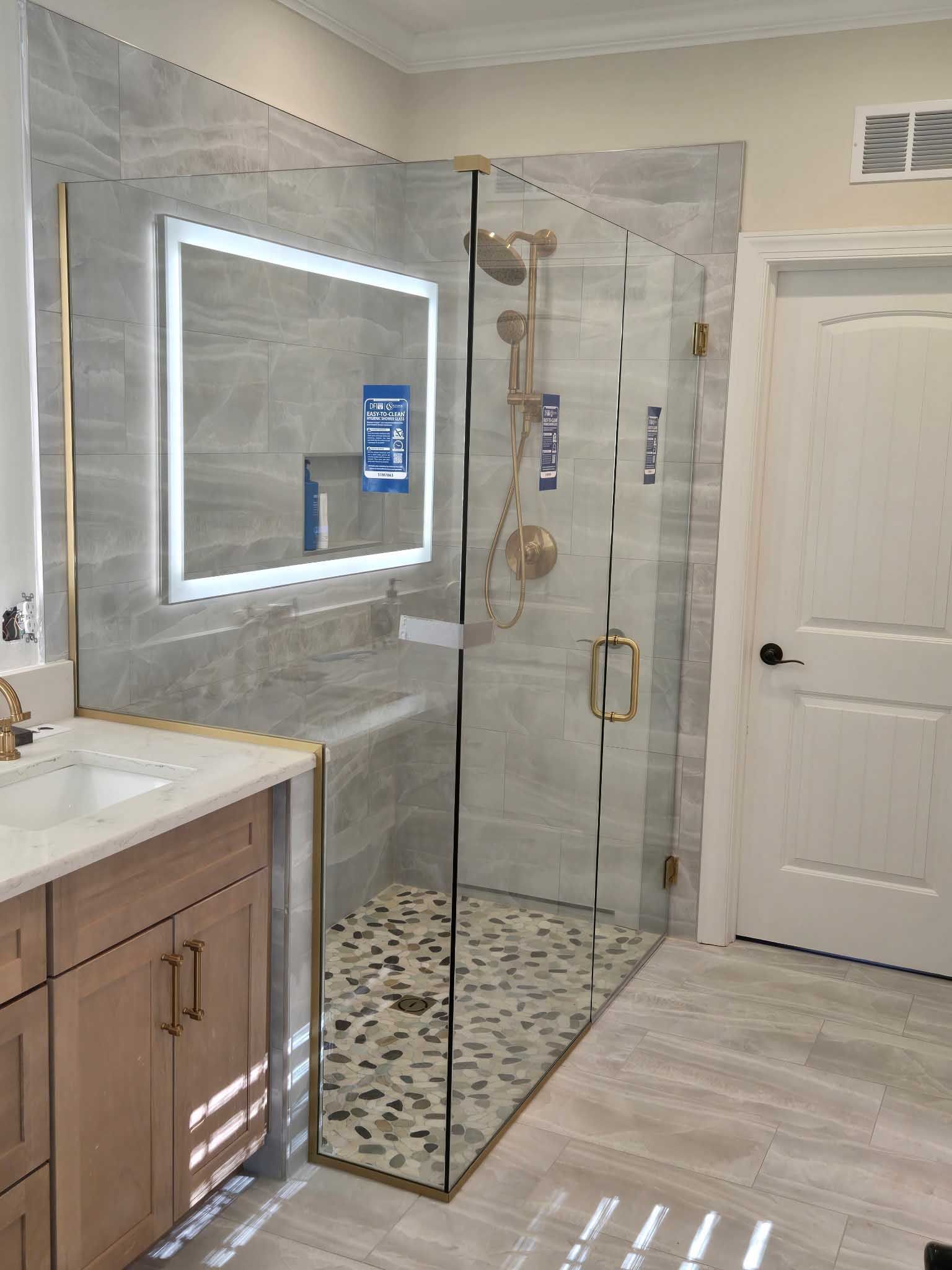 A modern bathroom with a wood vanity, a lighted mirror, and a glass-enclosed shower featuring gold fixtures and tile.
