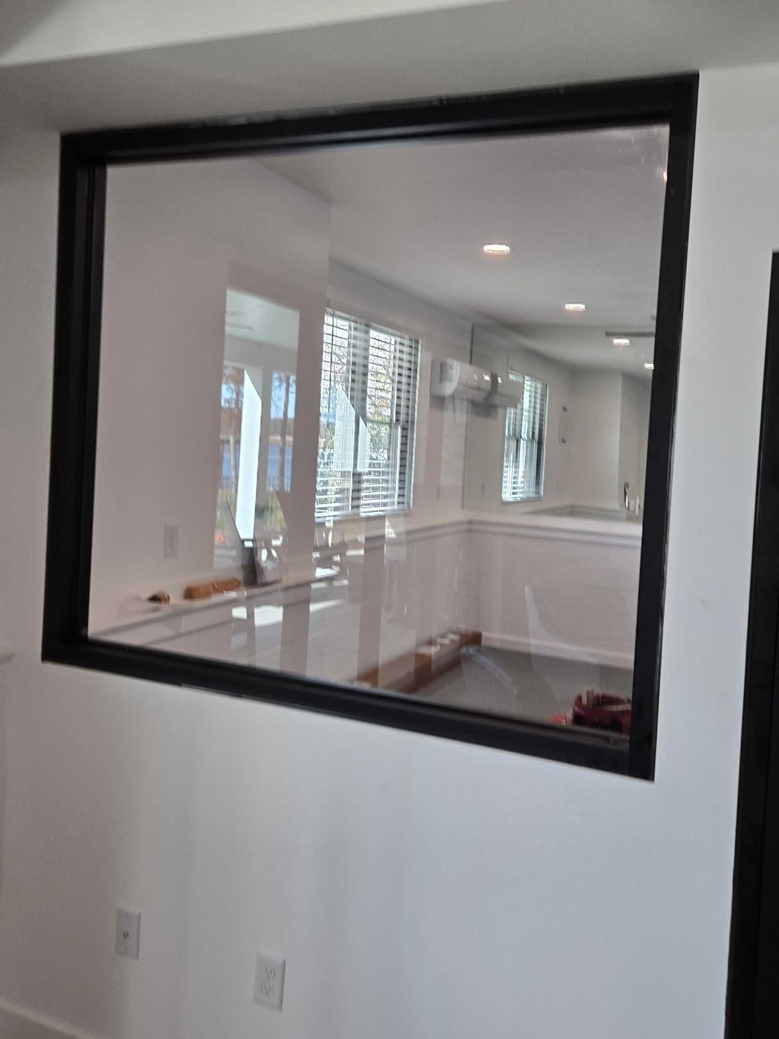 A large, rectangular indoor window with a dark frame set into a white wall, showing a bright, empty room reflected inside.