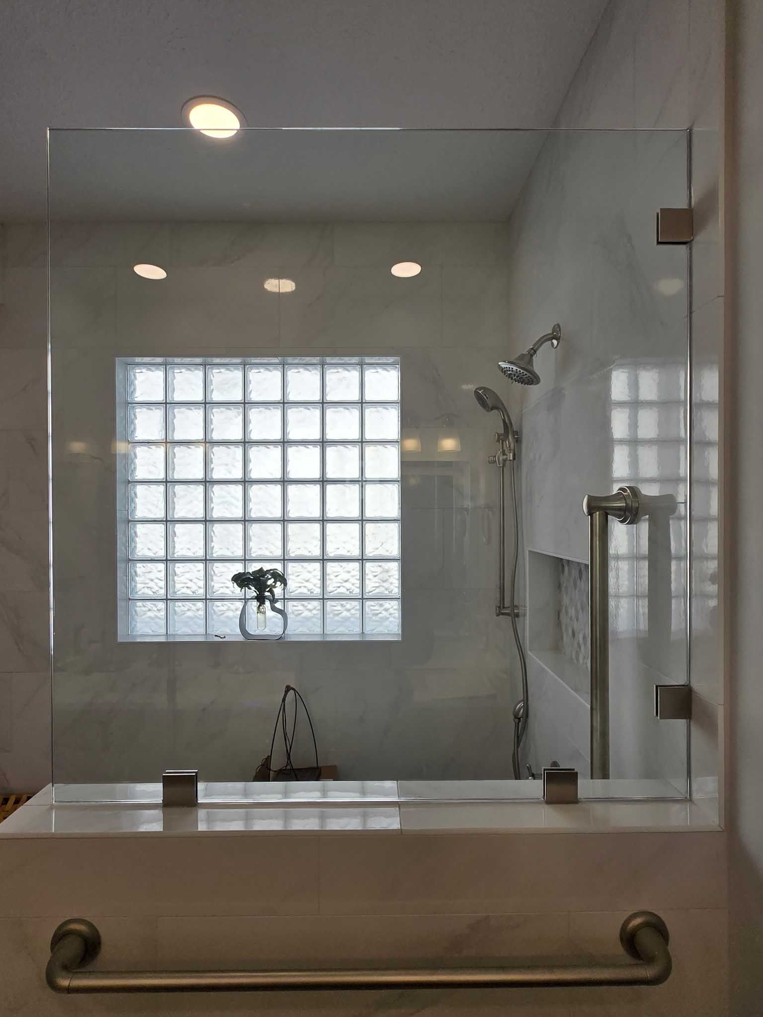 Glass shower partition with brushed metal hinges and a towel bar mounted on a marble ledge in a tiled bathroom.