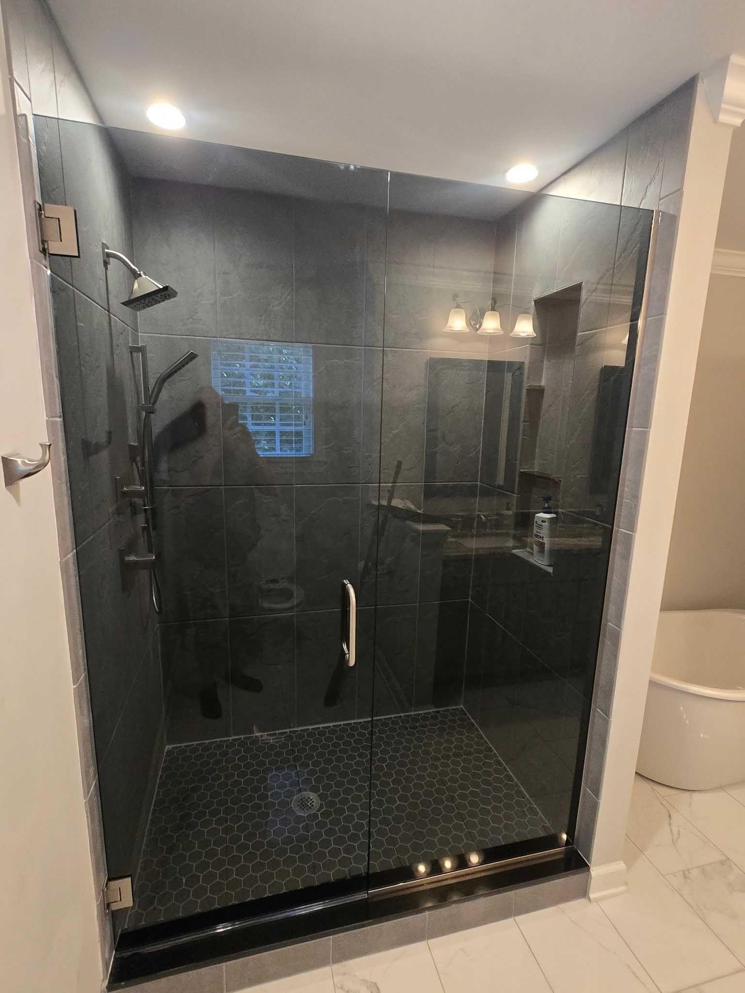 A modern walk-in shower with dark tiles, a glass door, a silver shower head, and a white freestanding tub nearby.