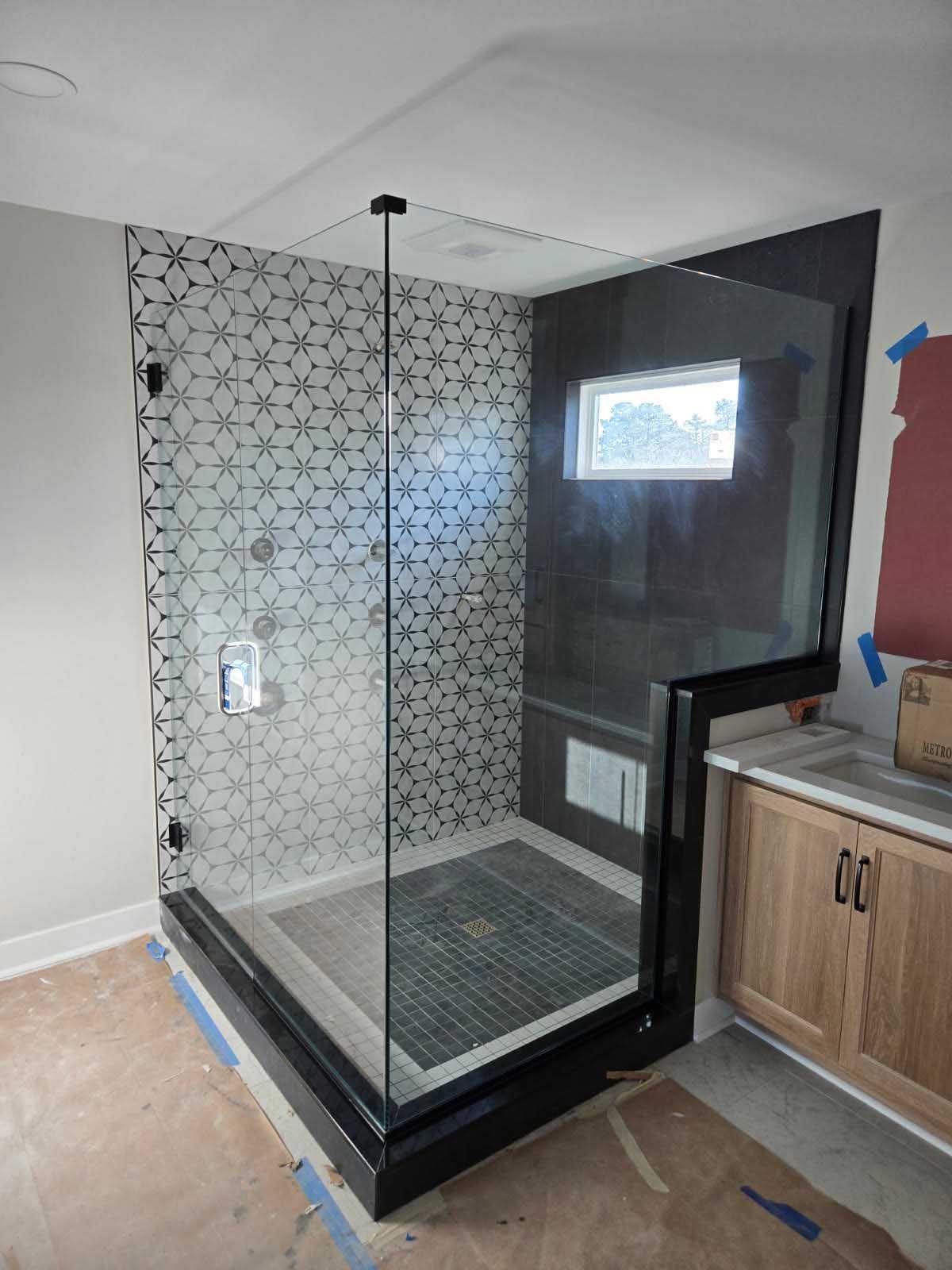 A bathroom under construction featuring a glass walk-in shower with black geometric tiles and a black marble curb.