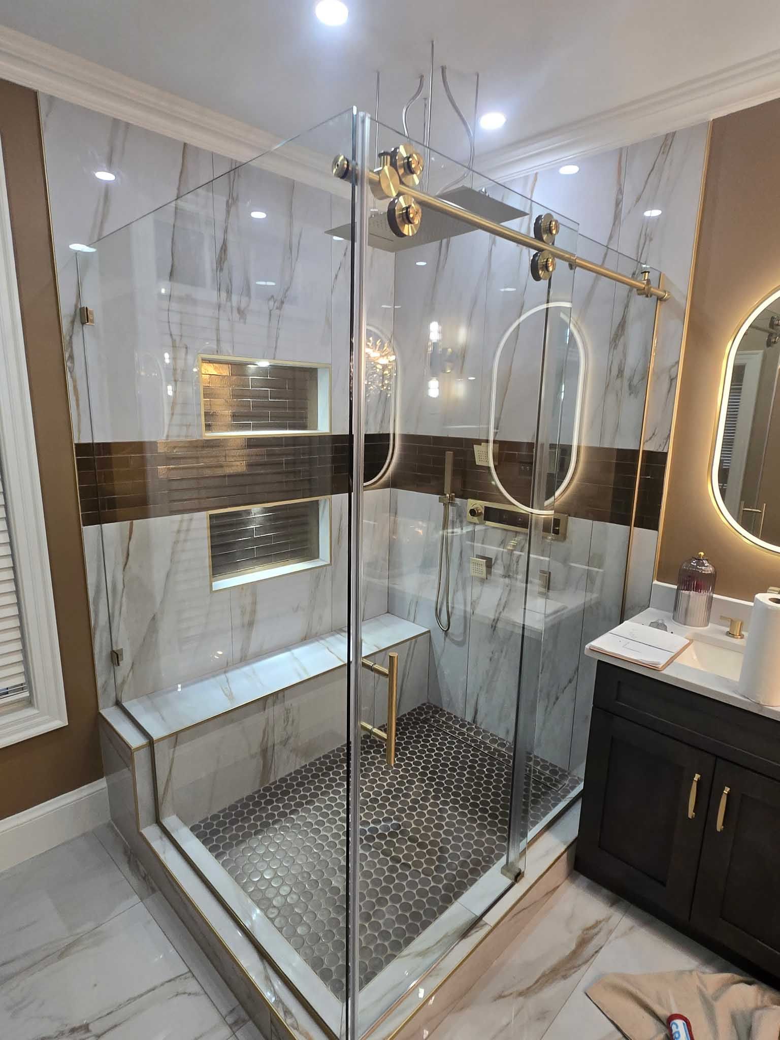 A walk-in shower with gold hardware, white marble-patterned walls, a brown accent tile, and a dark vanity with a mirror.