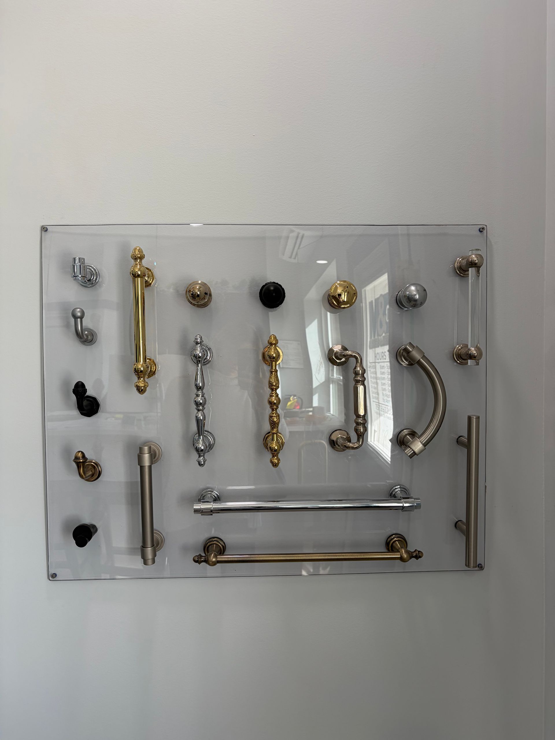 A clear acrylic wall display board showcasing an assortment of metal cabinet handles and knobs in various styles and finishes.