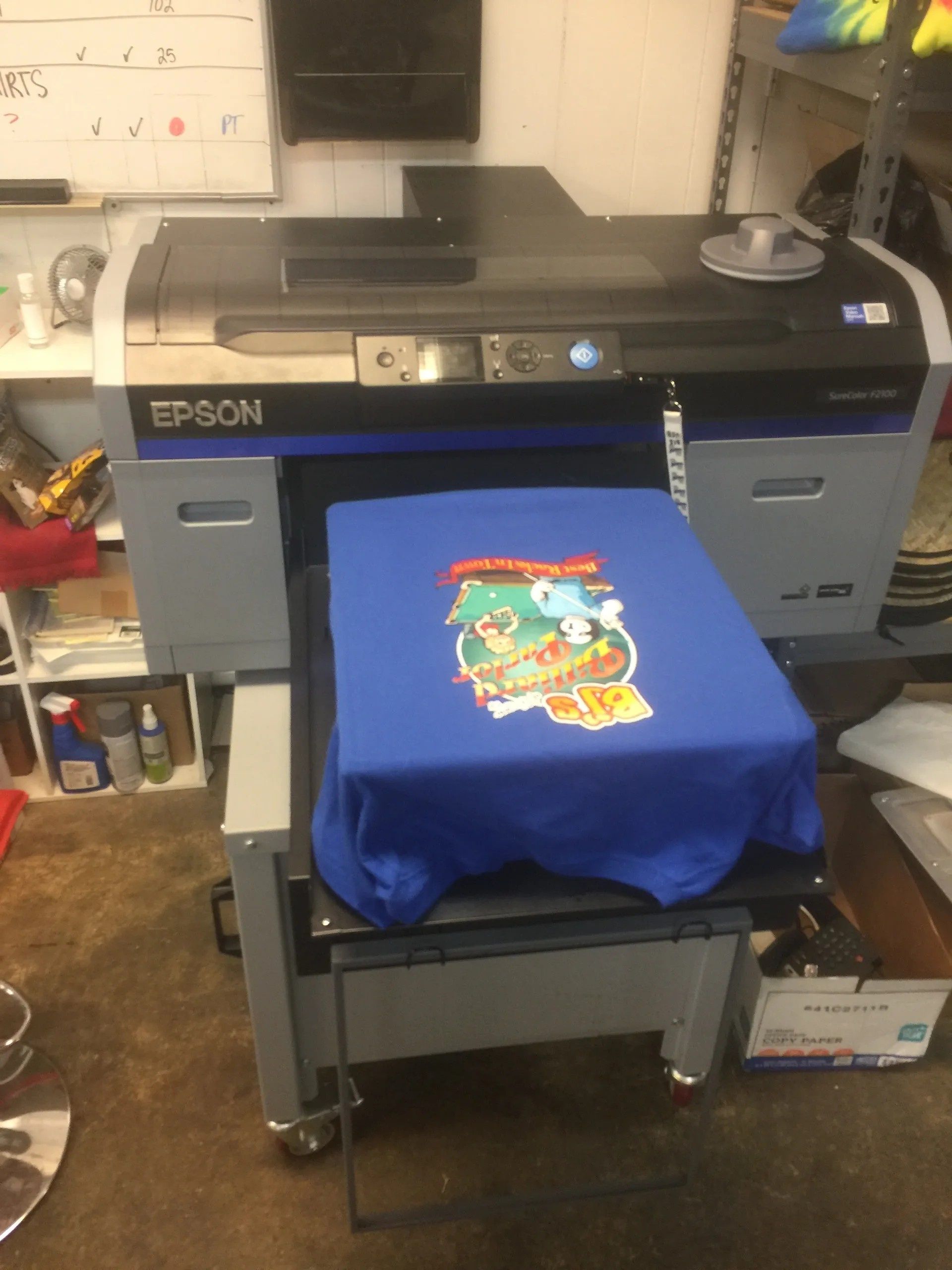 Epson printer printing a blue T-shirt with a colorful graphic in a workshop.