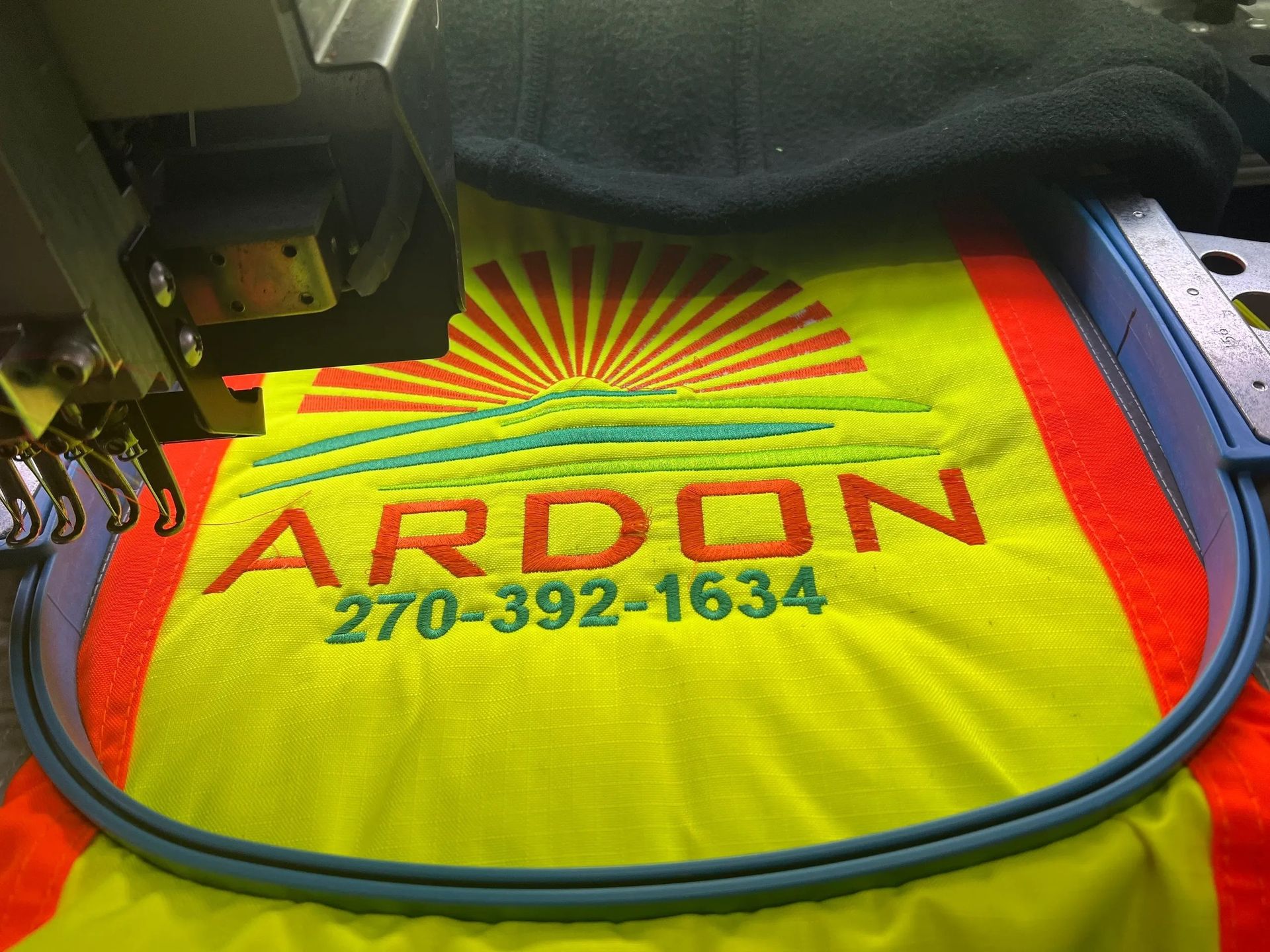 Embroidery machine stitching red ARDON logo and phone number on a yellow shirt with sunburst design