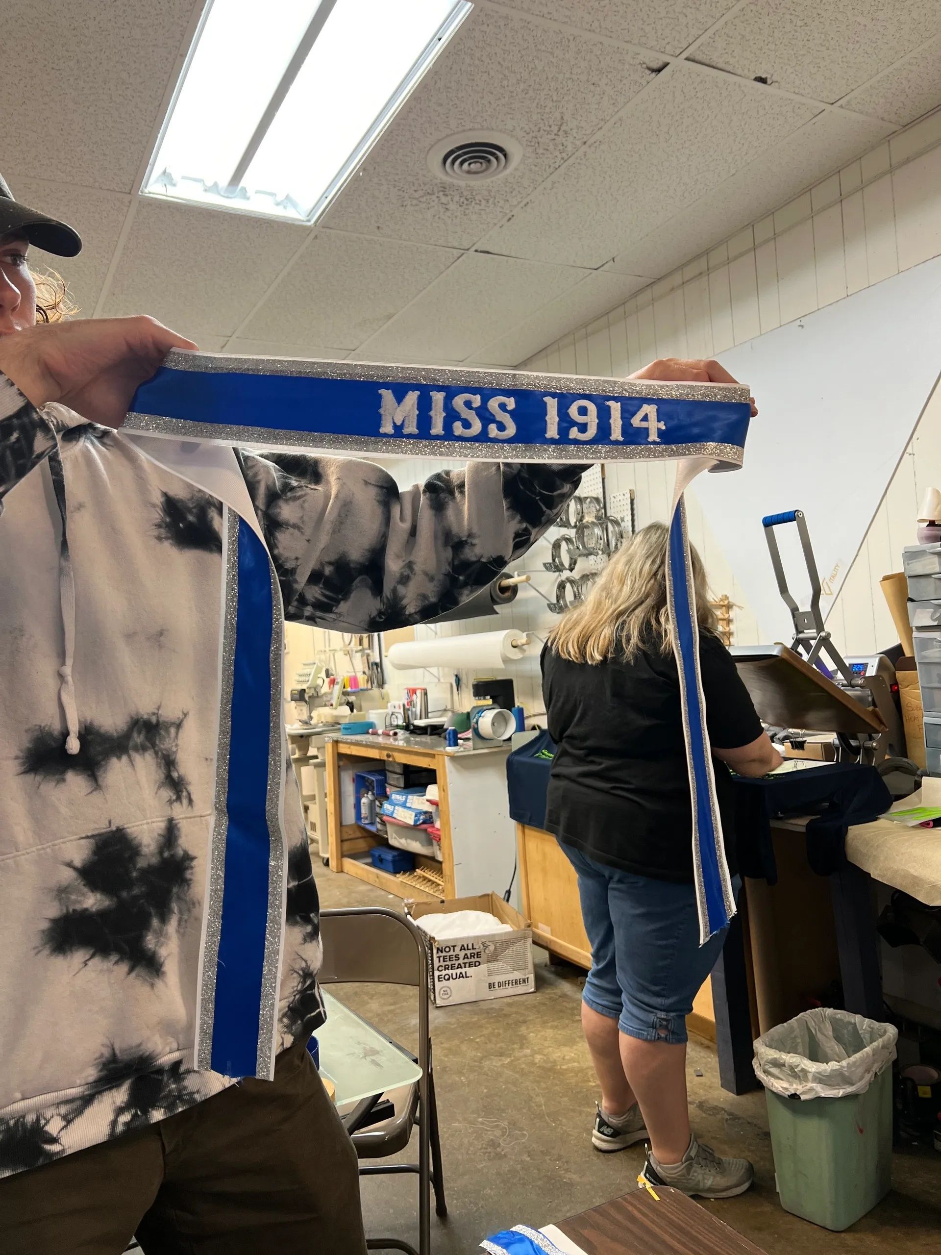 Blue “MISS 1914” sign held in a workshop, with two people and shelves in the background.