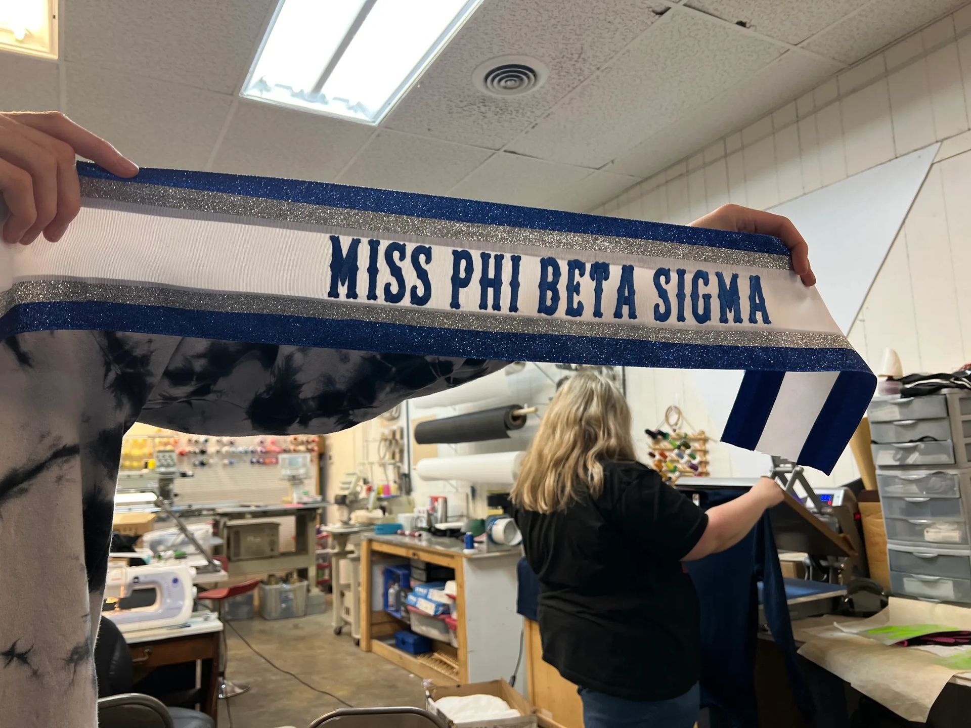 Blue-and-white “MISS PHI BETA SIGMA” banner held over a workshop scene