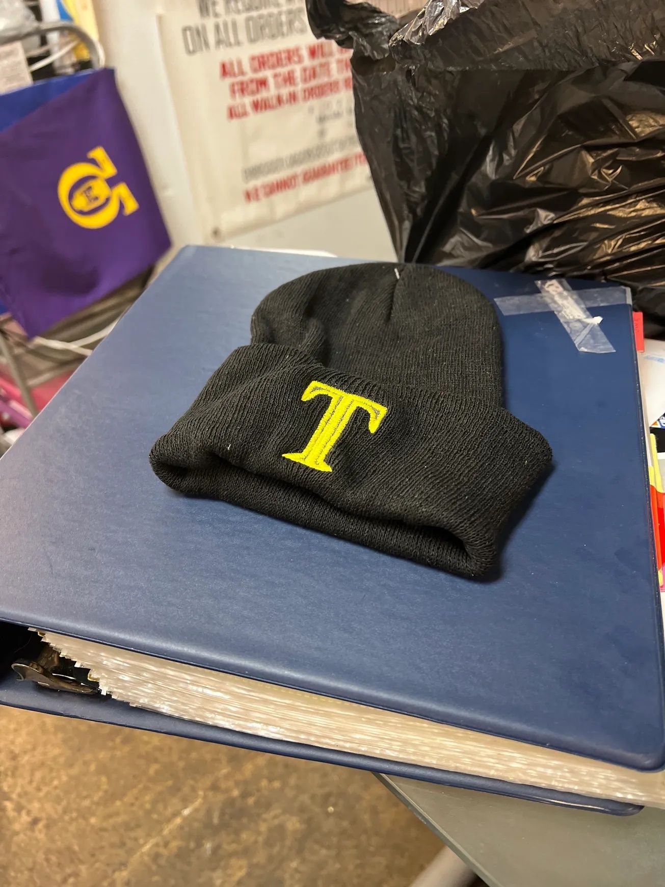 Black knit beanie with yellow T on a blue binder in a cluttered room
