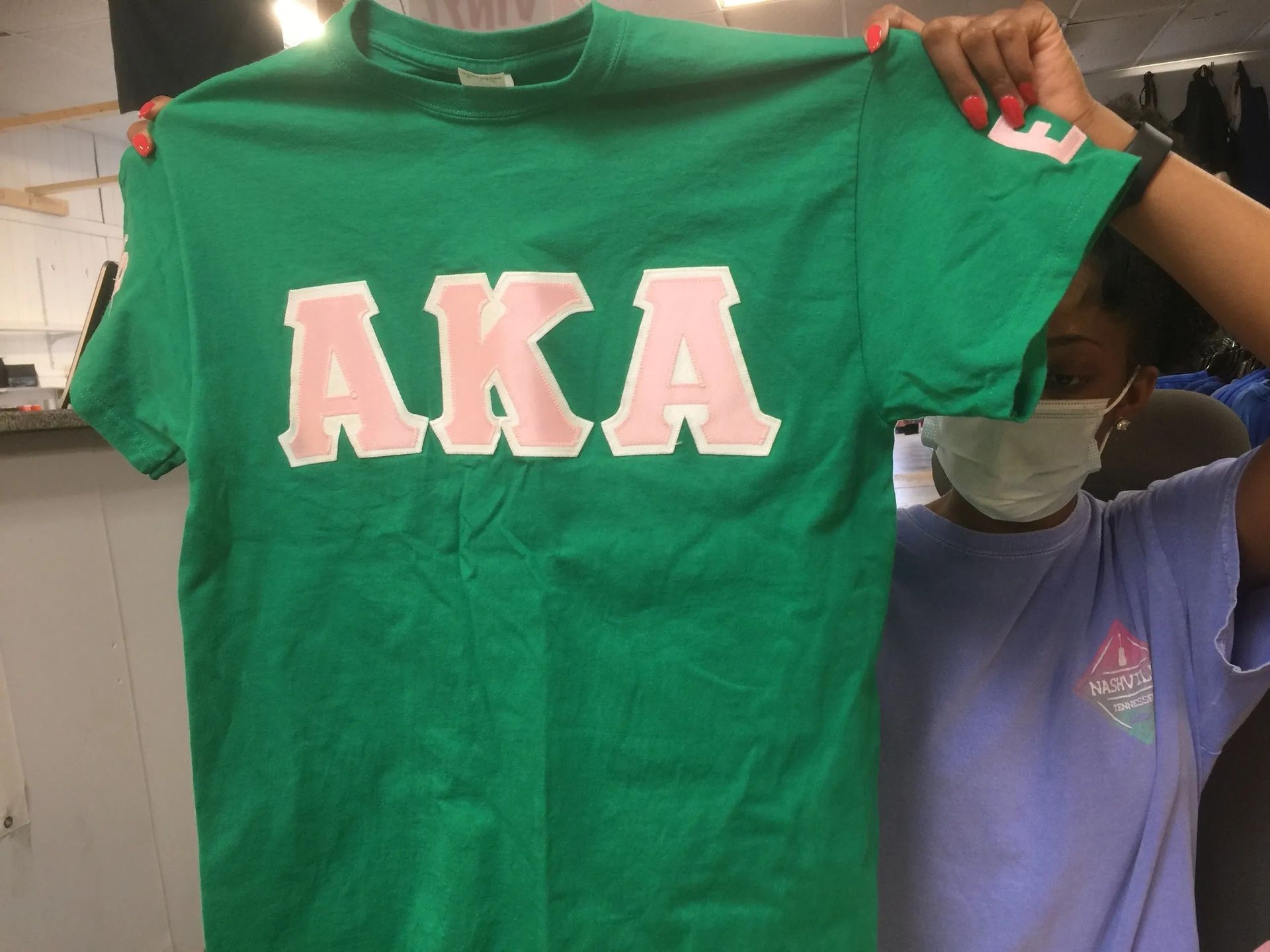 Green T-shirt with large pink “AKA” letters held up by a person indoors