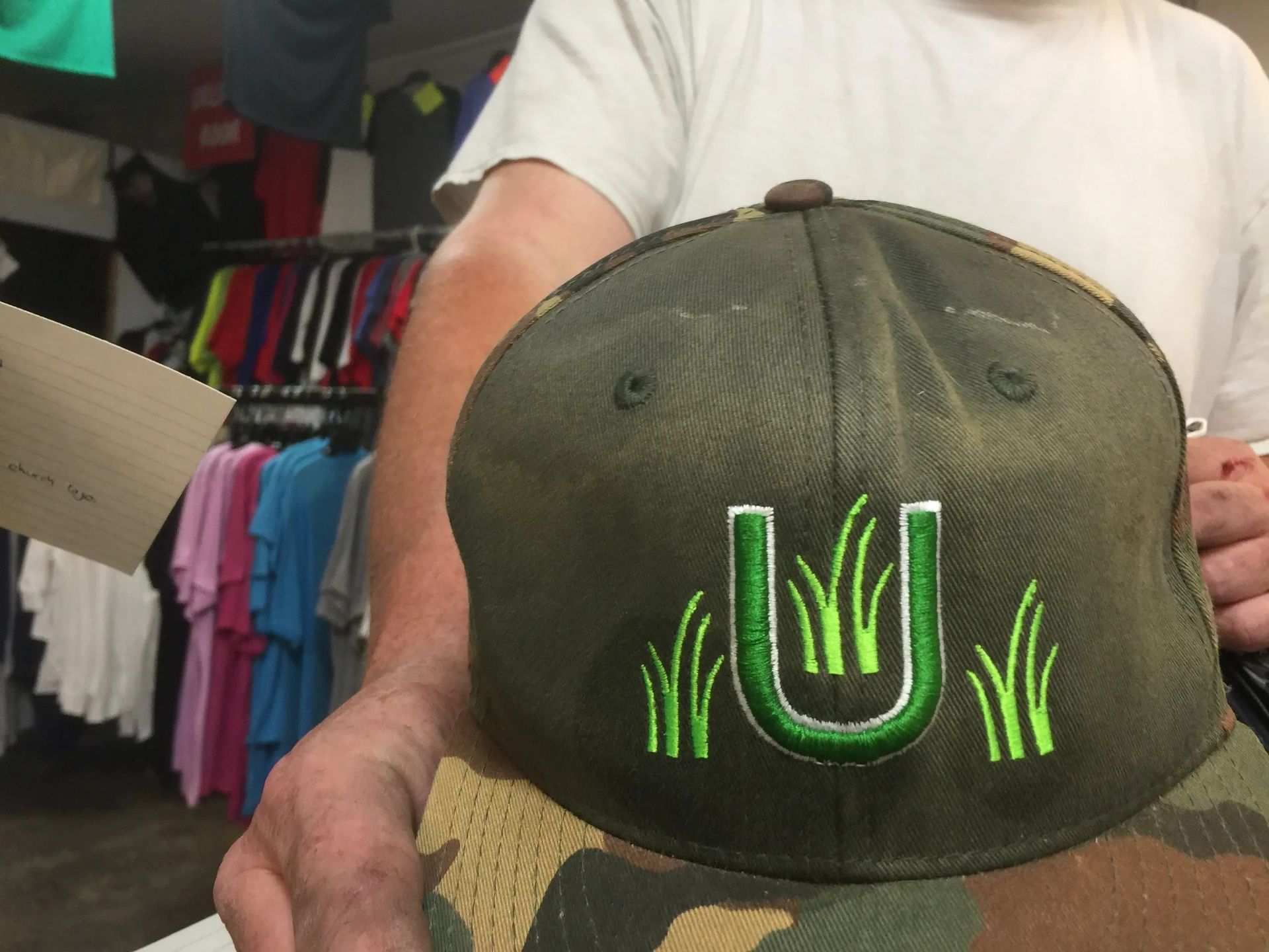 Olive green baseball cap with neon green embroidered design, held up in a clothing shop.