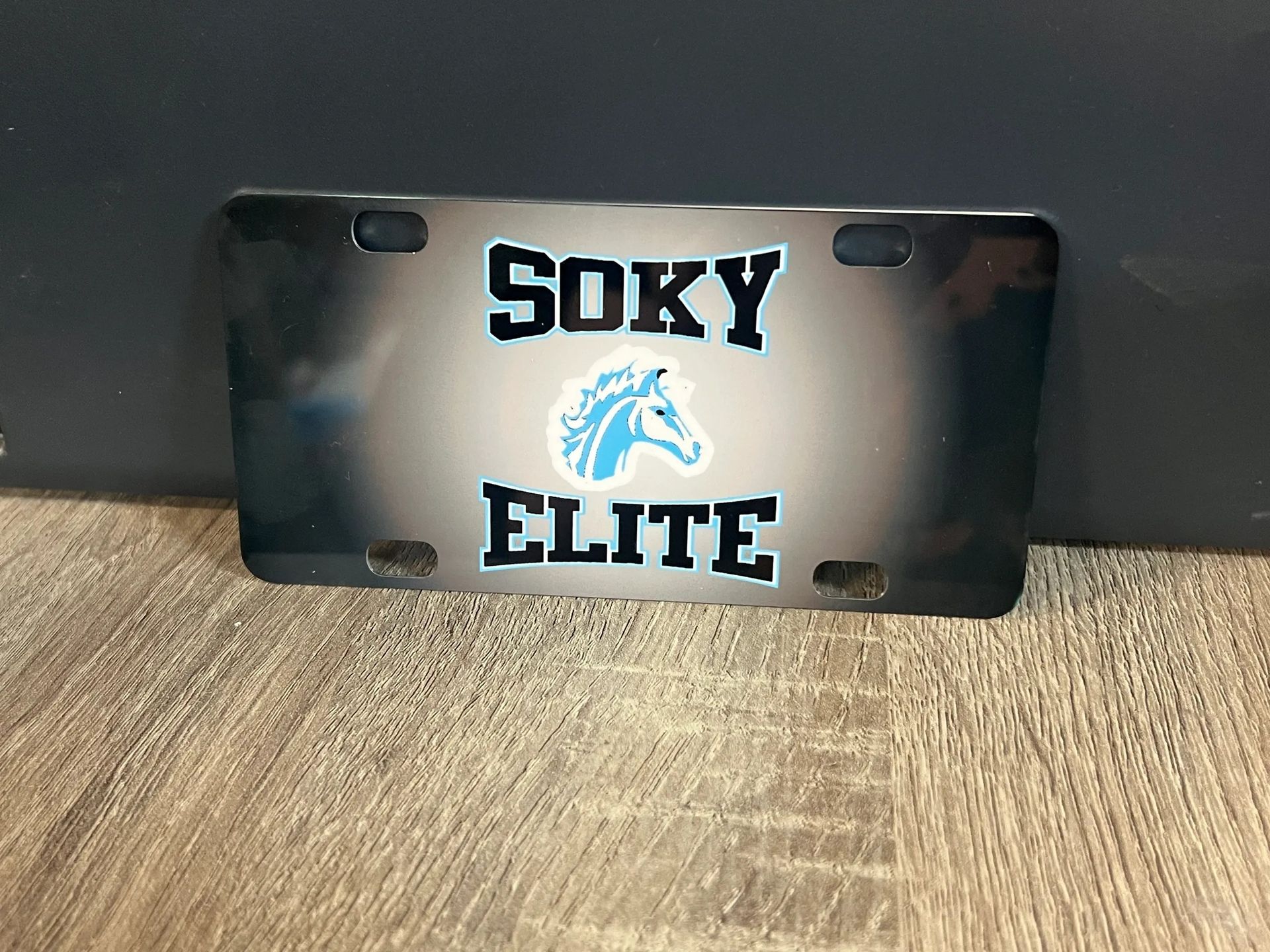 Black license plate with “SOKY ELITE” and a blue cougar logo on a wooden surface.