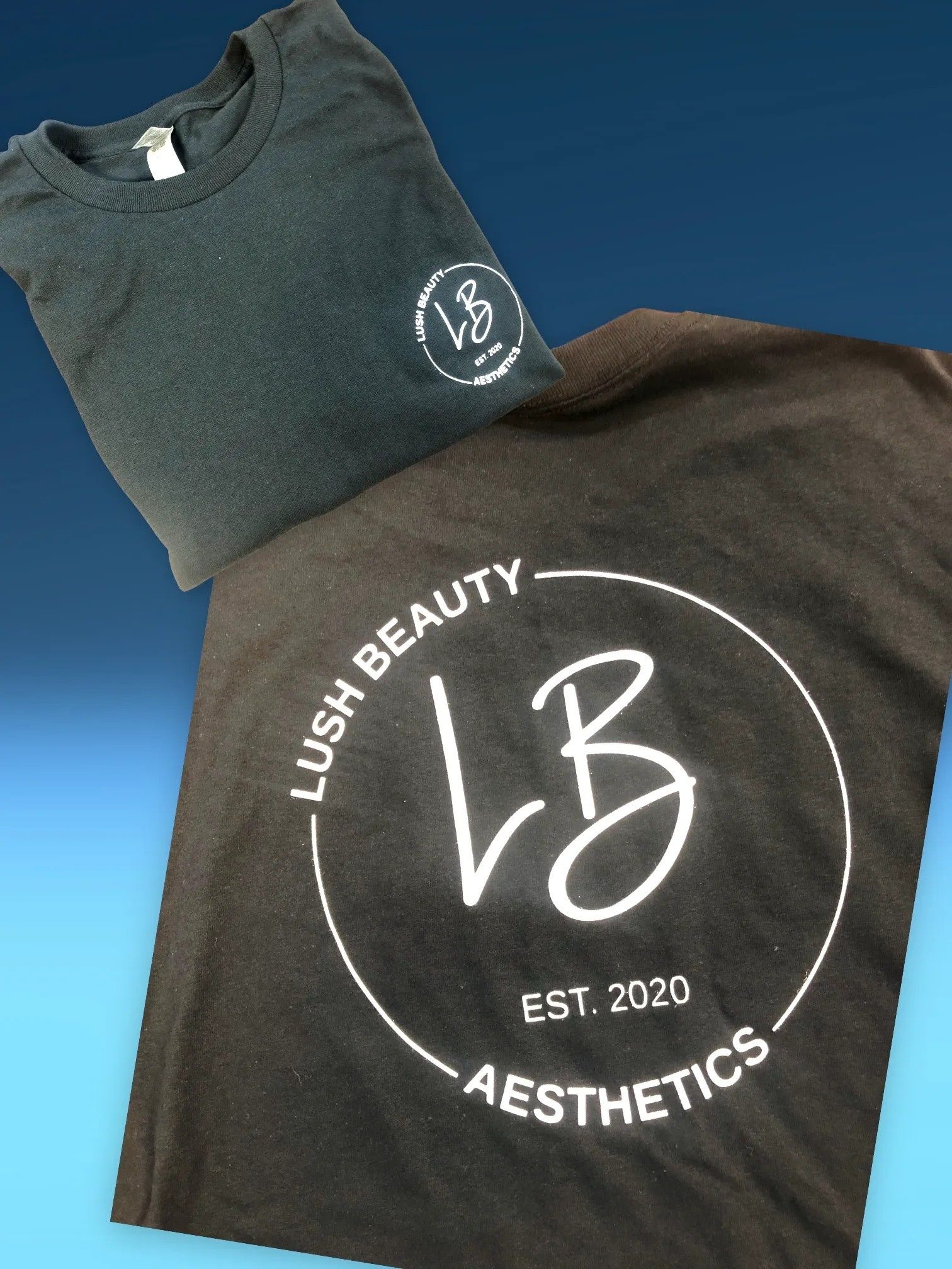 Two folded dark gray T-shirts with white Lush Beauty Aesthetics logos on a blue background