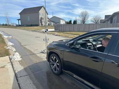 Black car parked on street next to a survey pole. A man is in the driver's seat on a sunny day.