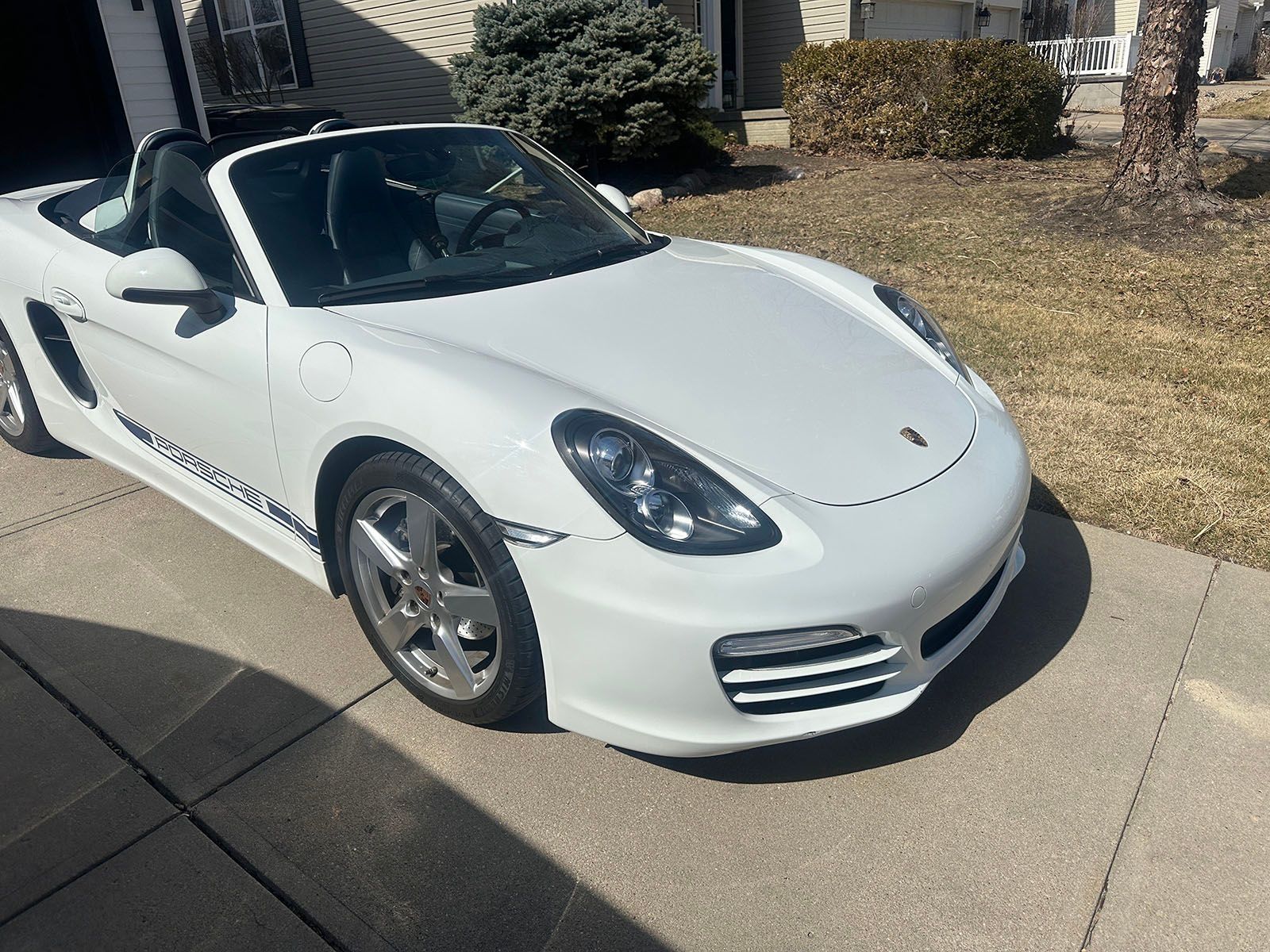 White Porsche Boxster convertible parked on a driveway in front of a house on a sunny day.