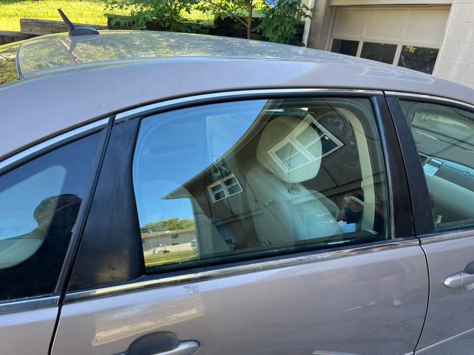 Silver car with person in driver's seat wearing protective face shield and clothing.