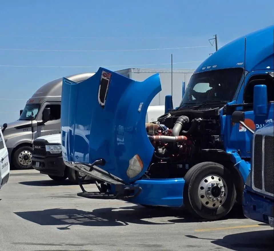 Blue semi-truck with its hood up, revealing the engine, parked in a lot. Gray and white trucks in the background.