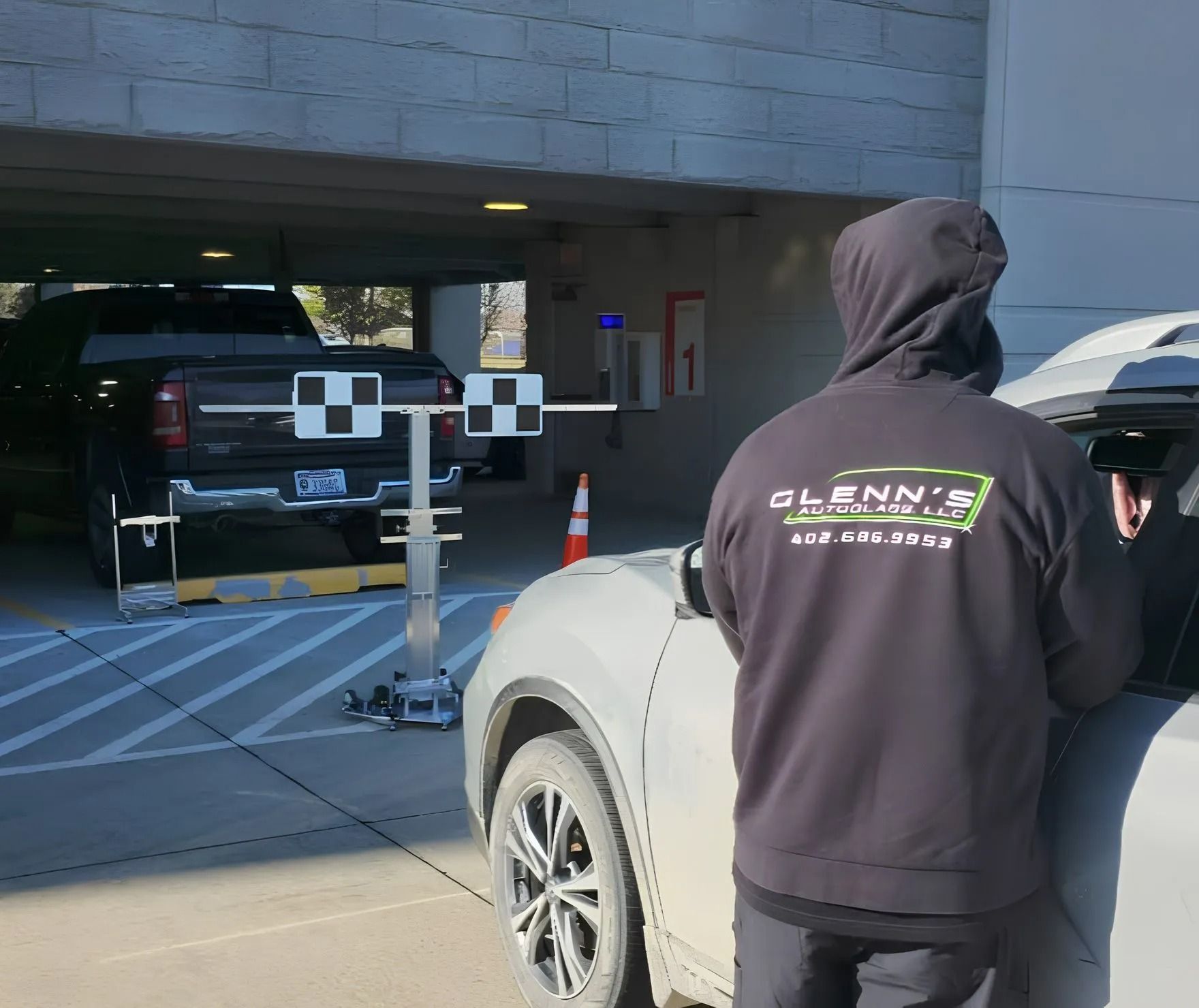 Person in a hooded sweatshirt performing car alignment at a garage. Alignment equipment is set up, pickup truck in background.