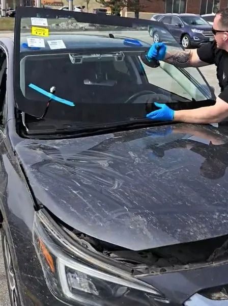 Person replacing a car windshield. A damaged gray car has a new windshield being installed outside.