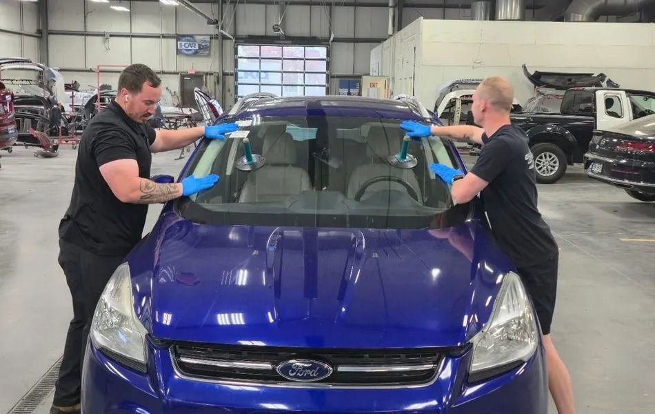 Two men installing a windshield on a blue Ford SUV in a garage.