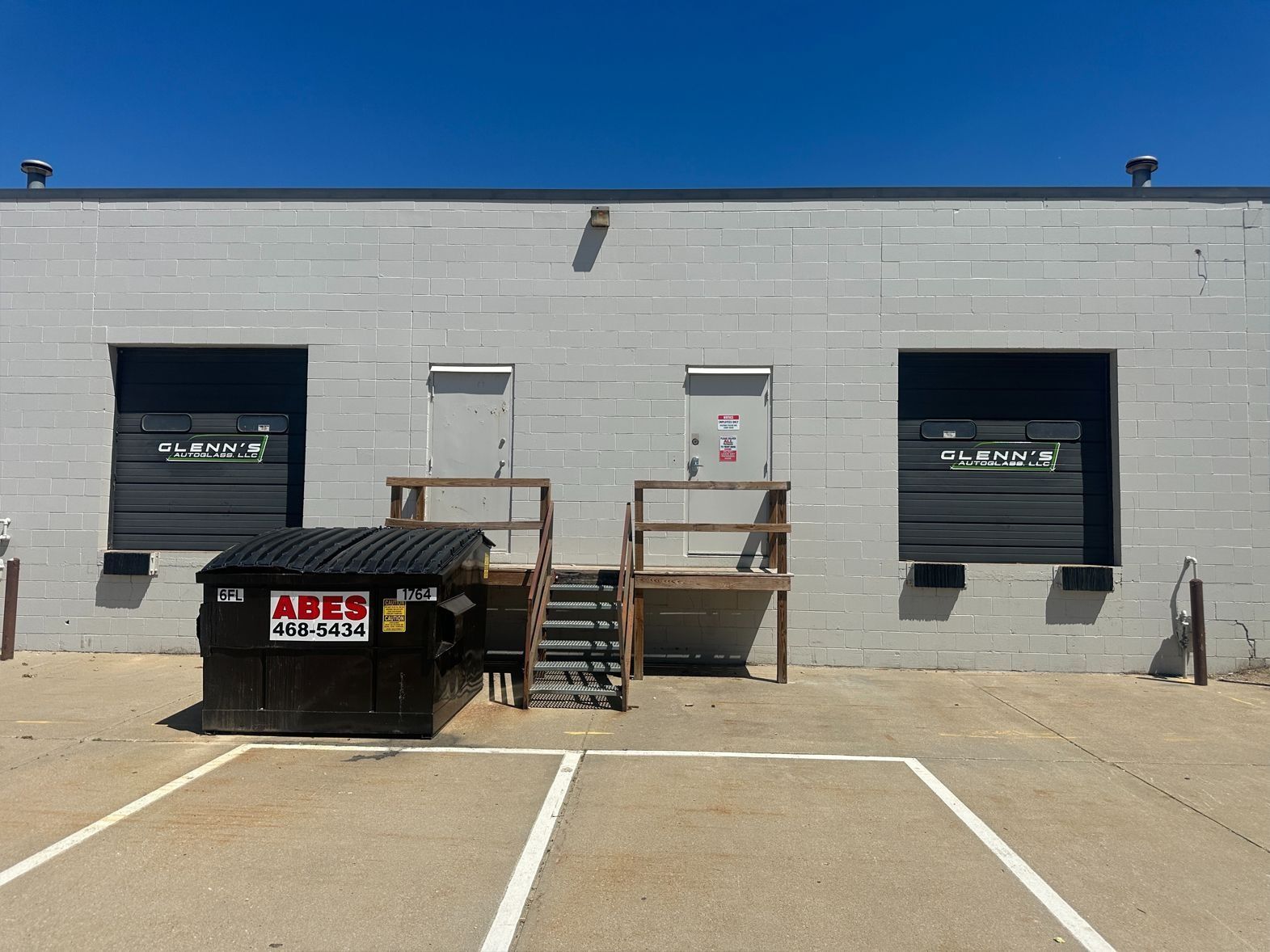 Exterior of a gray building with loading docks, steps, a dumpster, and marked parking spaces under a clear blue sky.
