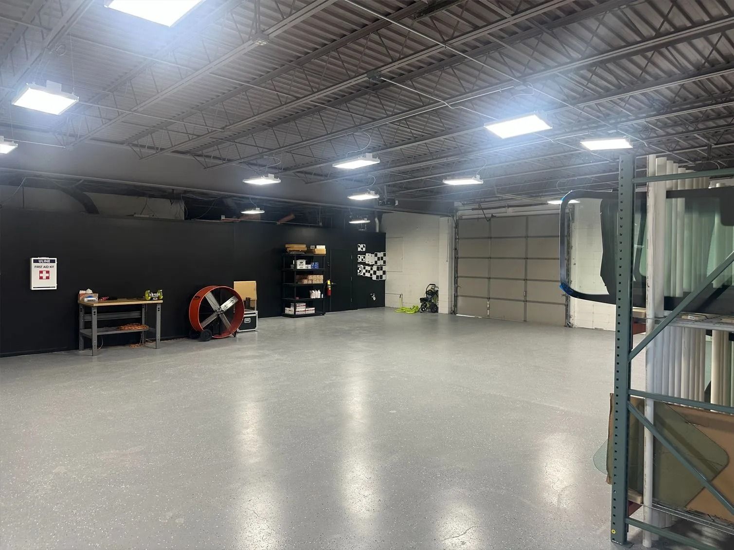 Spacious warehouse with gray floor, black wall, shelves, fan, and overhead lighting.
