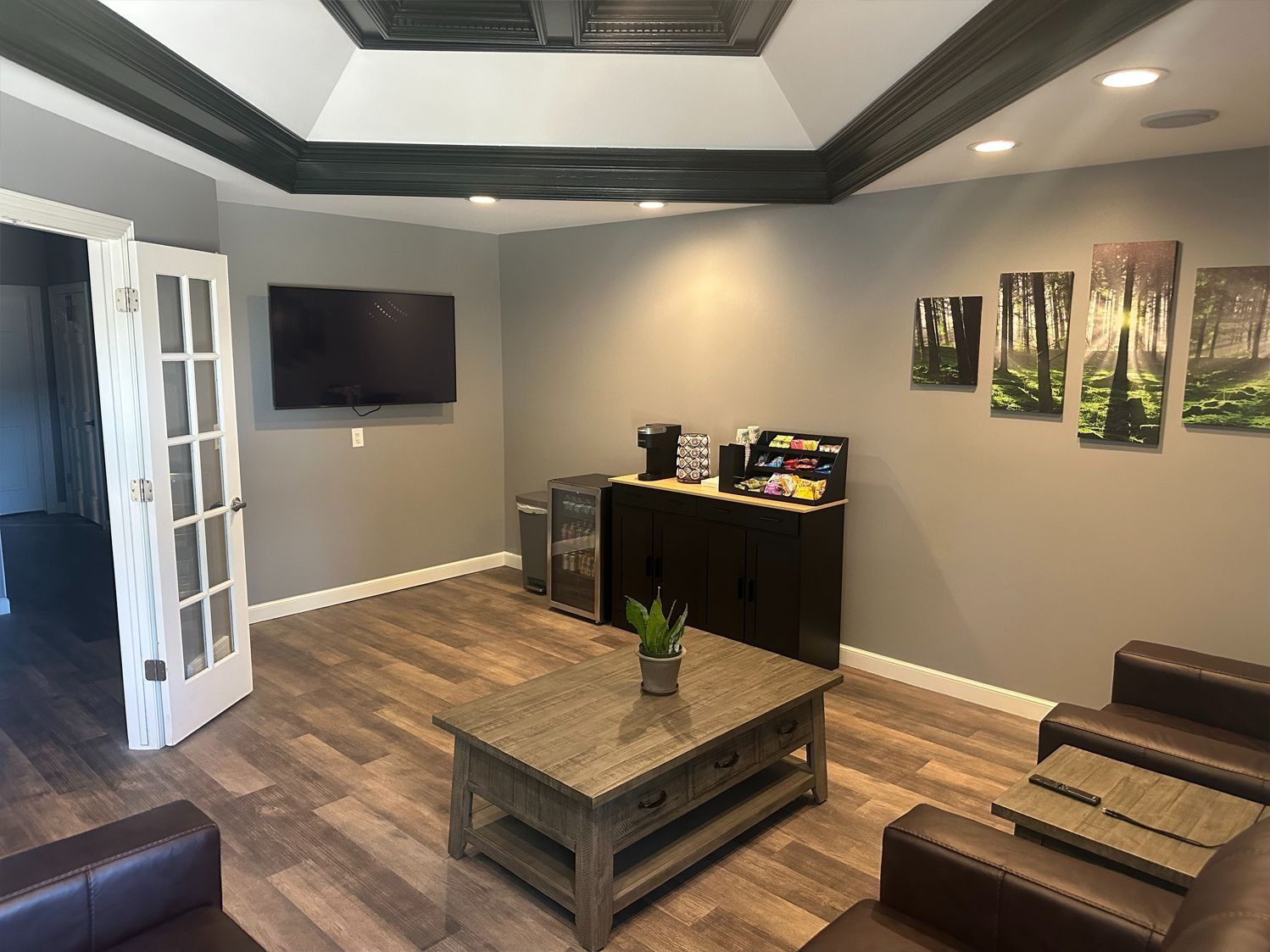 Lounge area with brown leather seating, coffee table, TV, and refreshments. Gray walls and dark trim, wood-look flooring.