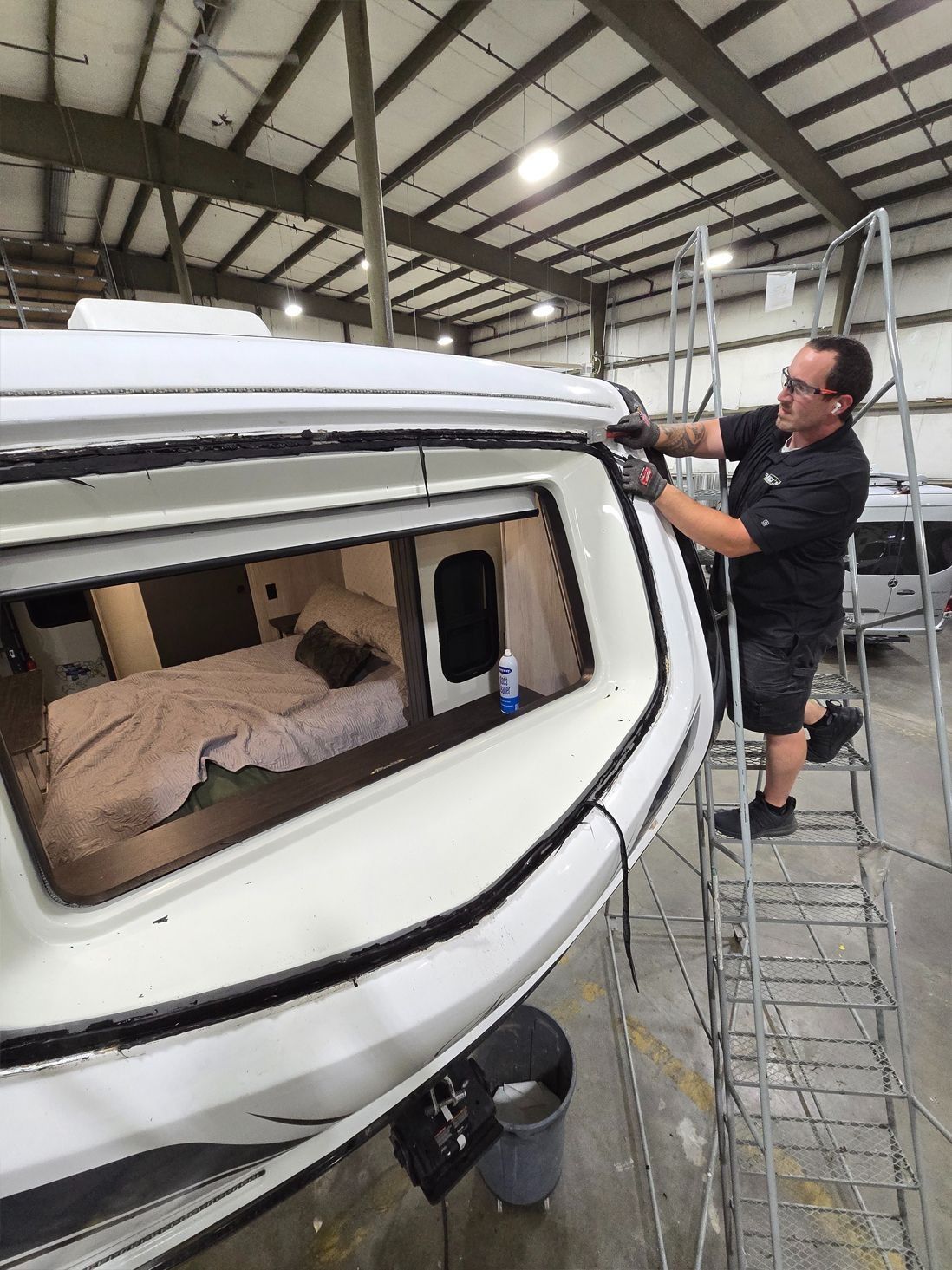 Man on ladder installing trim on a white camper shell. Inside, a bed is visible. Shop setting.