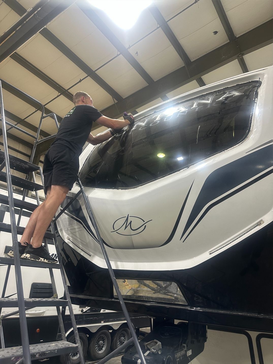 Person applying tint to a large vehicle window, working from a ladder, inside a shop.