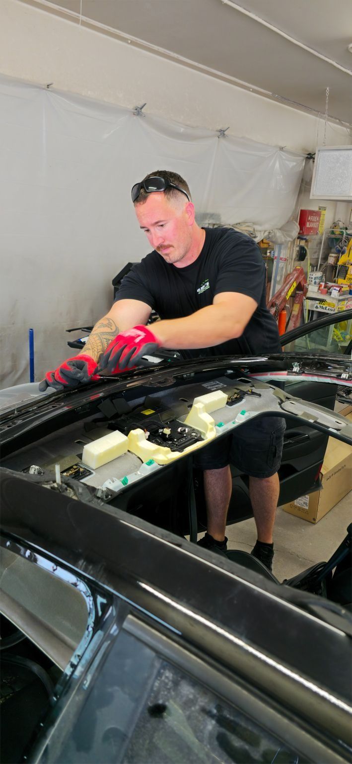 A man with gloves works on a car roof in a garage. He wears a black shirt and shorts.
