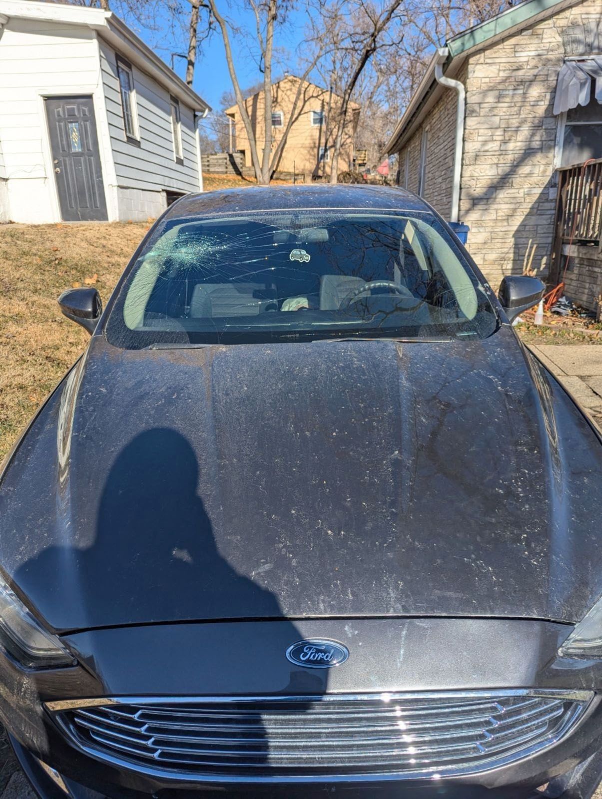 A dark gray Ford car covered in dust parked outdoors on a sunny day, with small buildings in the background.