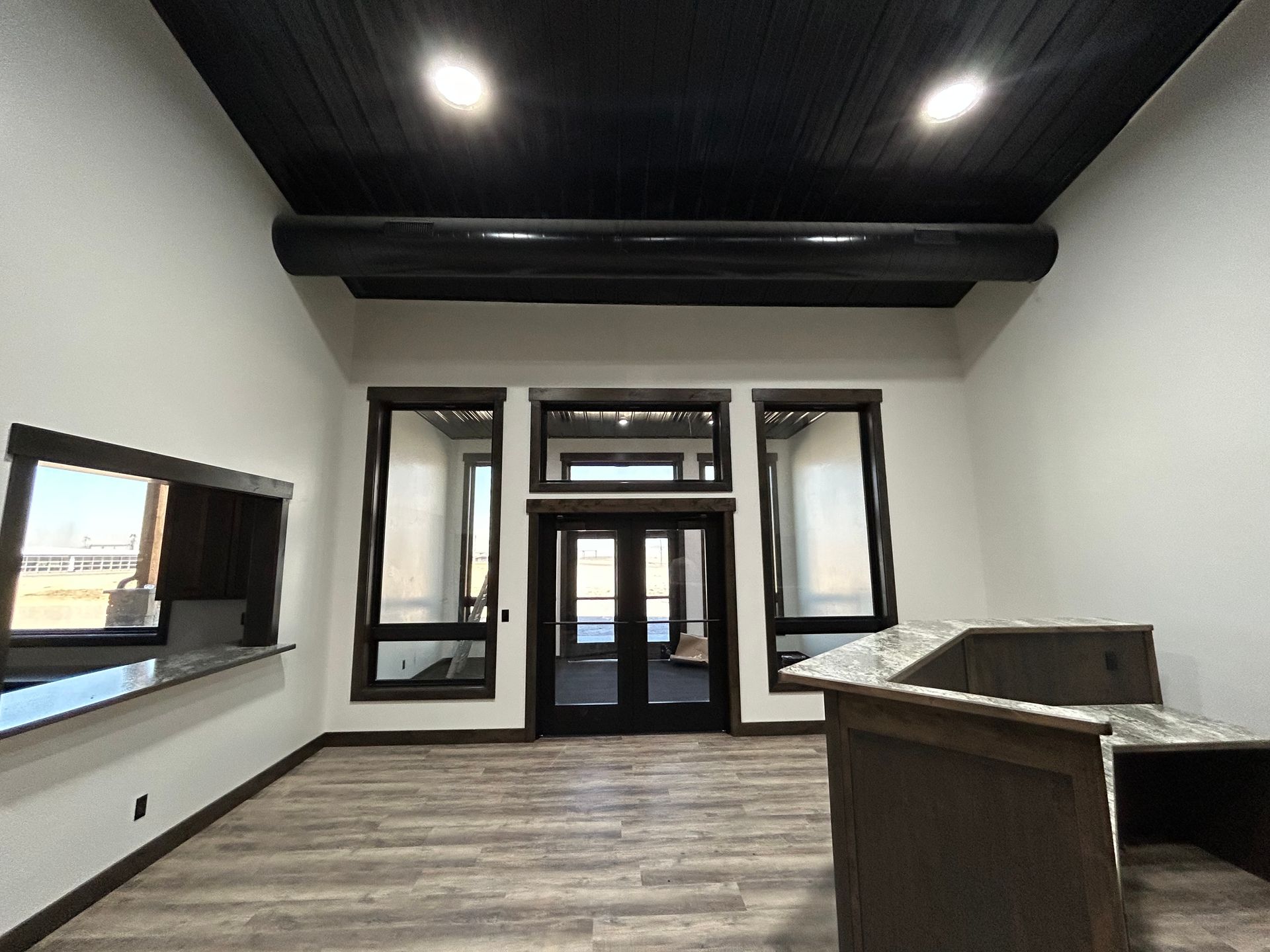 Empty modern room with black ceiling beams, wood floor, large windows, and a front entry door.