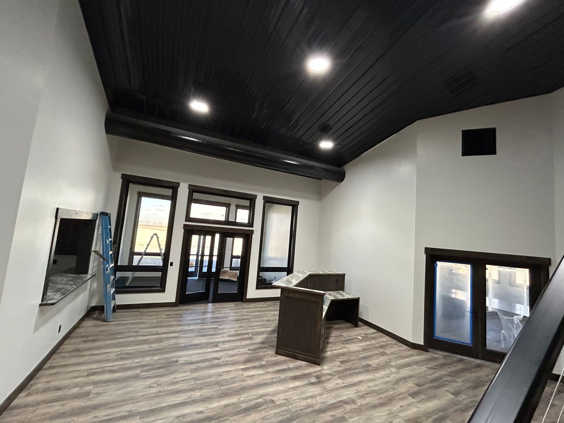 Empty modern interior with white walls, dark ceiling, wood-look floor, and windows/doors at the far end.