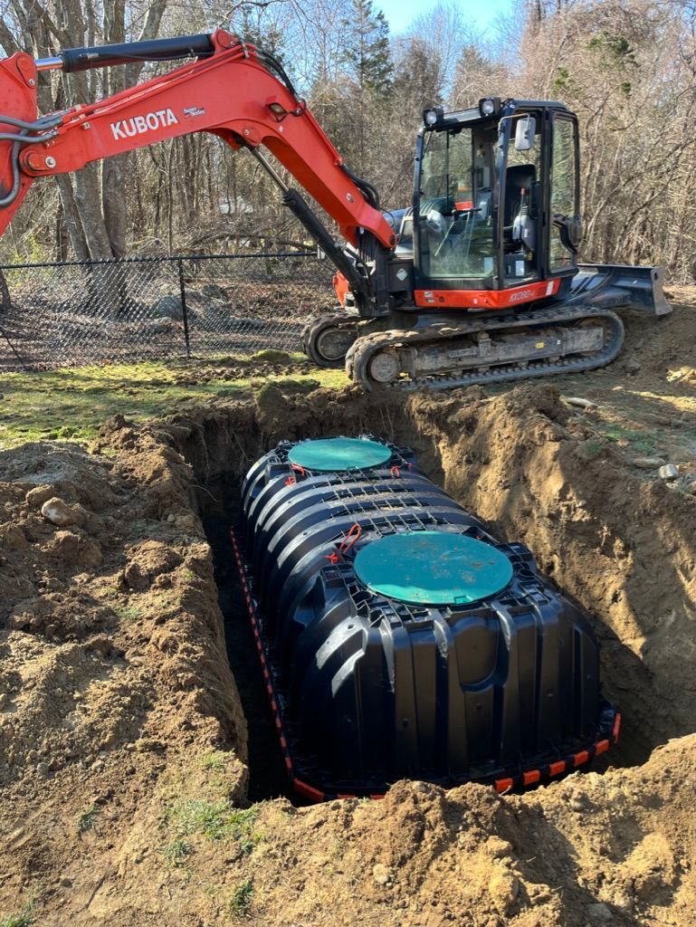 A black septic tank sits in an excavated trench as an orange Kubota excavator hovers above.