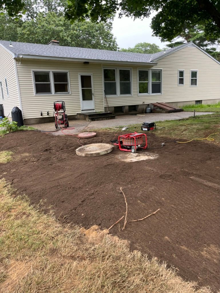 Backyard with freshly tilled soil, a house in the background, and construction equipment on a patio.