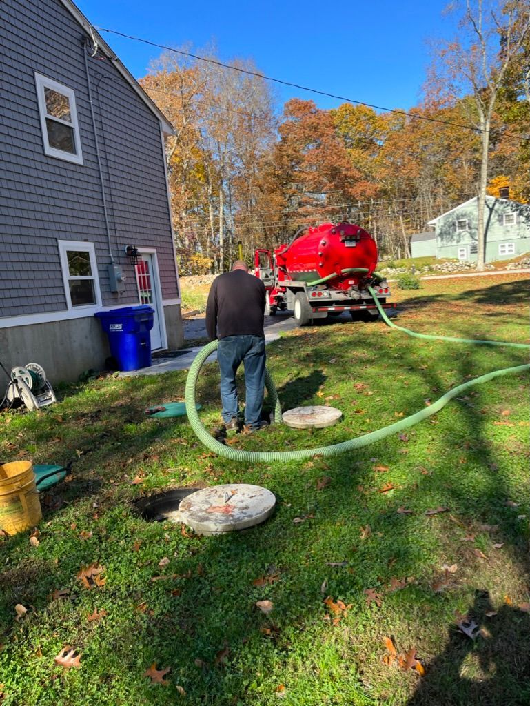 Man pumping septic tank in a yard with a red truck, next to a house under a clear sky.
