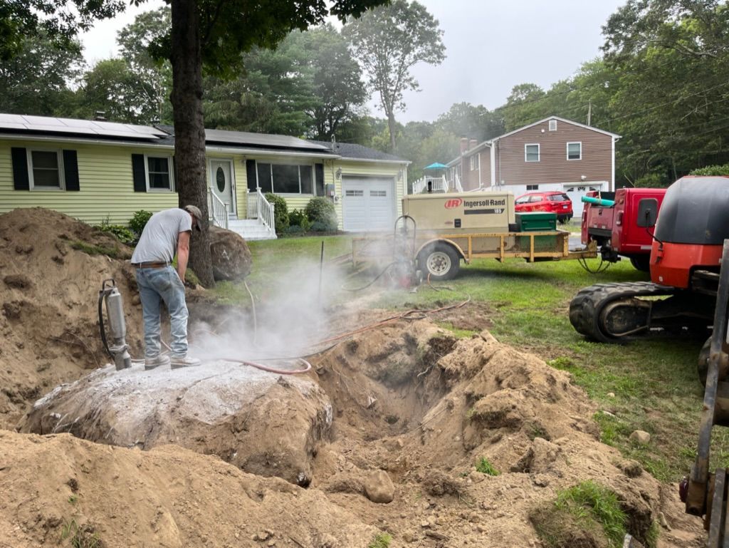 Man using jackhammer on rock in yard; construction equipment and homes in background.