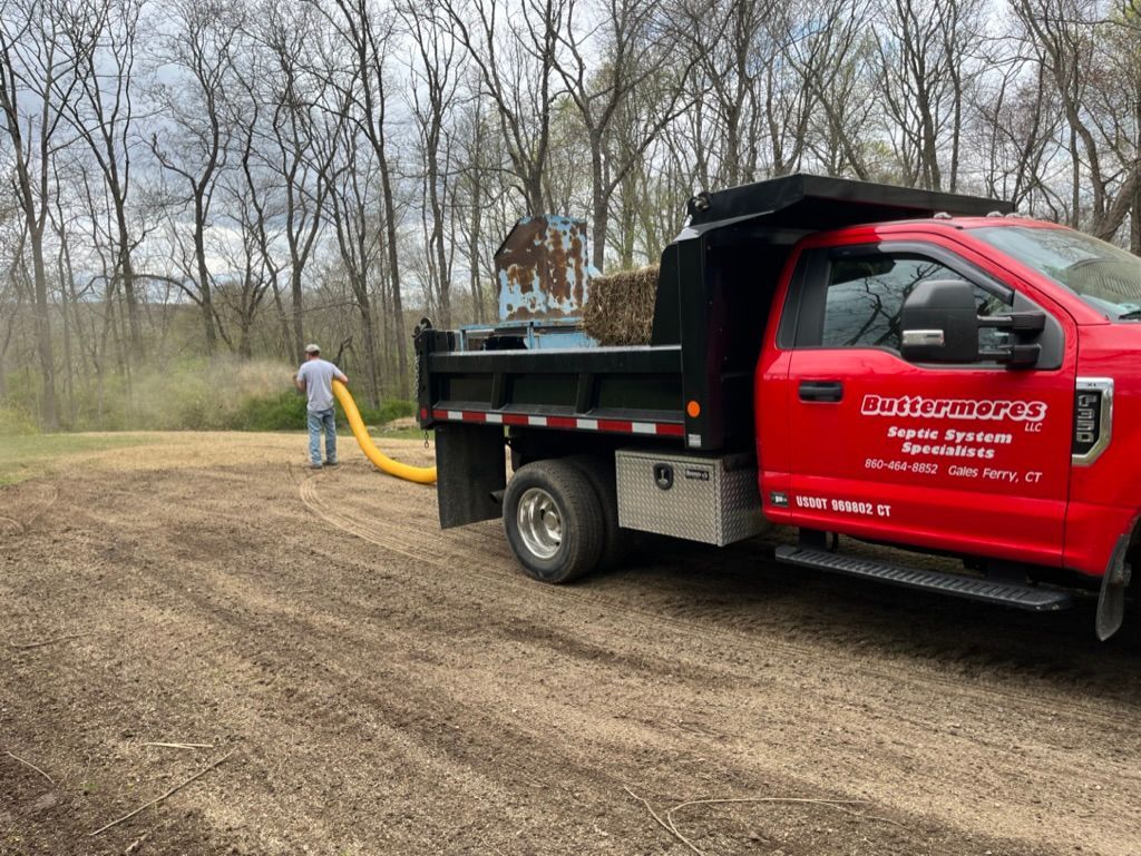 Red dump truck with a person using a hose to distribute material onto the ground near trees.