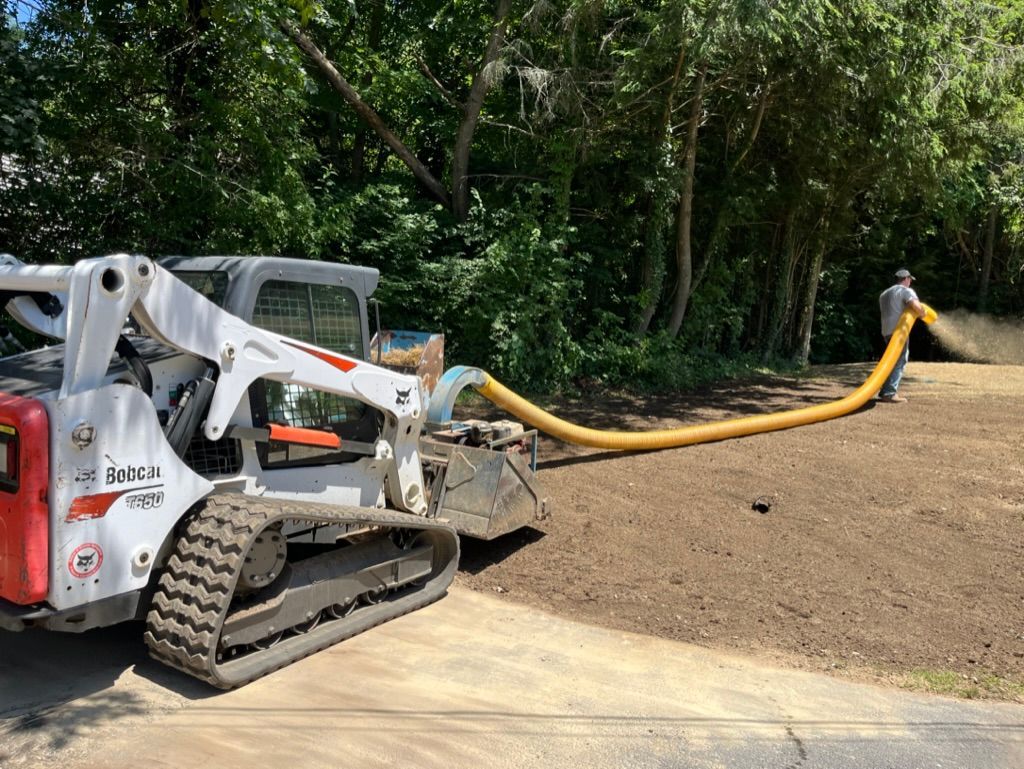 Bobcat skid steer sprays mulch onto a lawn, worker holds hose.