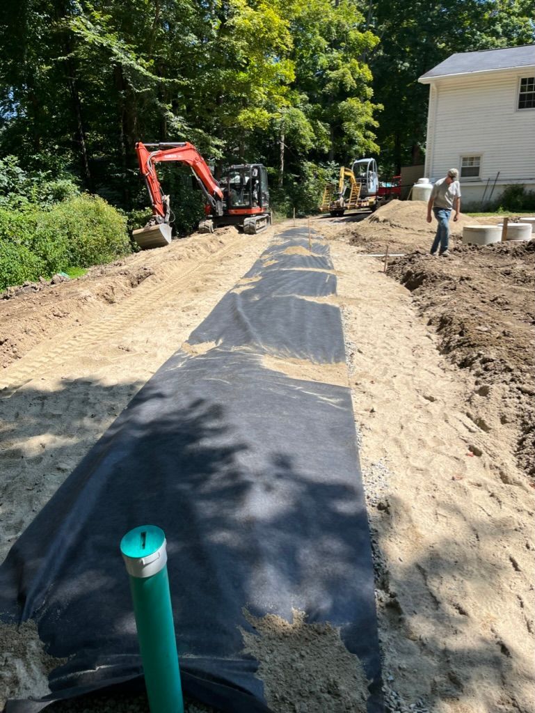 Construction site: long trench with black fabric. Excavators and worker visible.