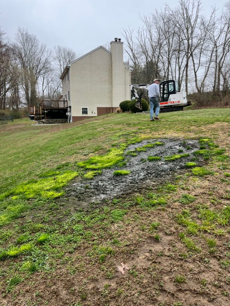 Man walking toward a Bobcat excavator on a grassy hillside near a house with a deck.