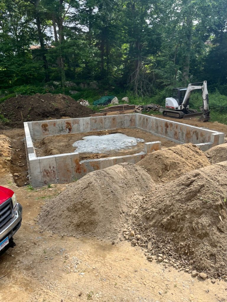 Foundation of a building under construction, surrounded by dirt piles and an excavator, in a wooded area.