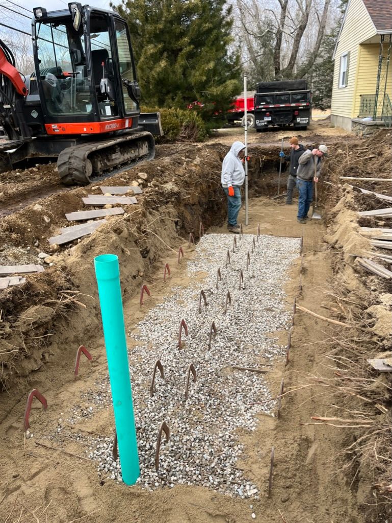 Construction site with workers, excavator, and truck; trench with gravel and pipes.