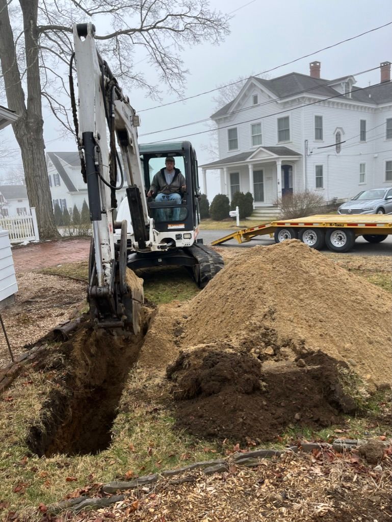 Excavator digging a trench on a residential lawn, with operator visible. Pile of dirt beside the trench and house in background.