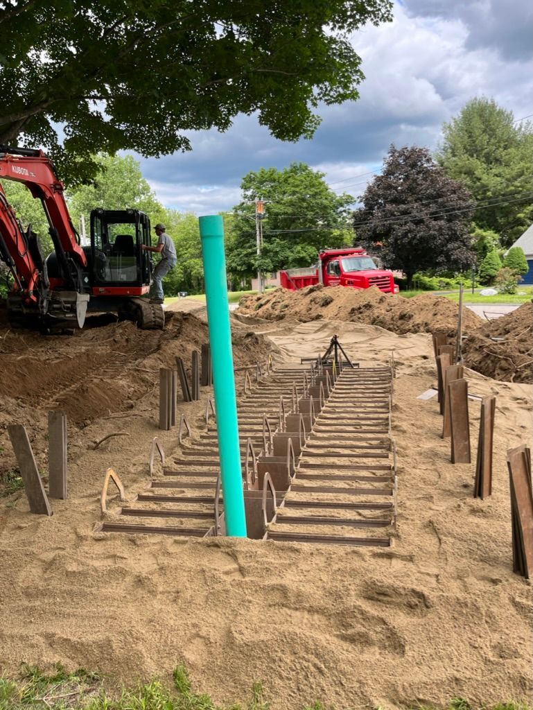 Construction site with an excavator, trench, and a green pipe. Worker near the back. Red truck in background. Cloudy day.