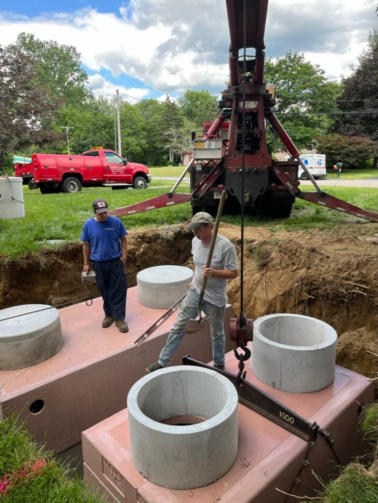 Workers installing a septic system. A crane lowers concrete rings onto brown tanks in an excavated area. Red truck in the background.