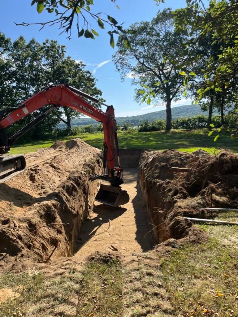 An excavator digging a trench in a grassy area, with trees and a distant landscape in the background.